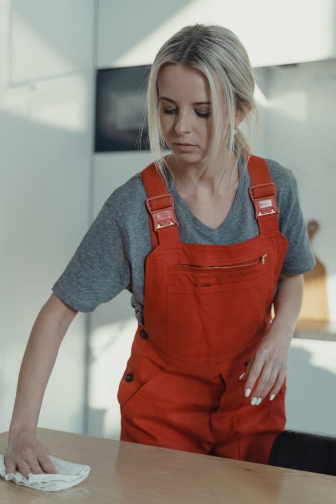 pexels-photo-6195109 Blonde woman in red overalls sanitizing a wooden table indoors, showcasing home cleaning service.
